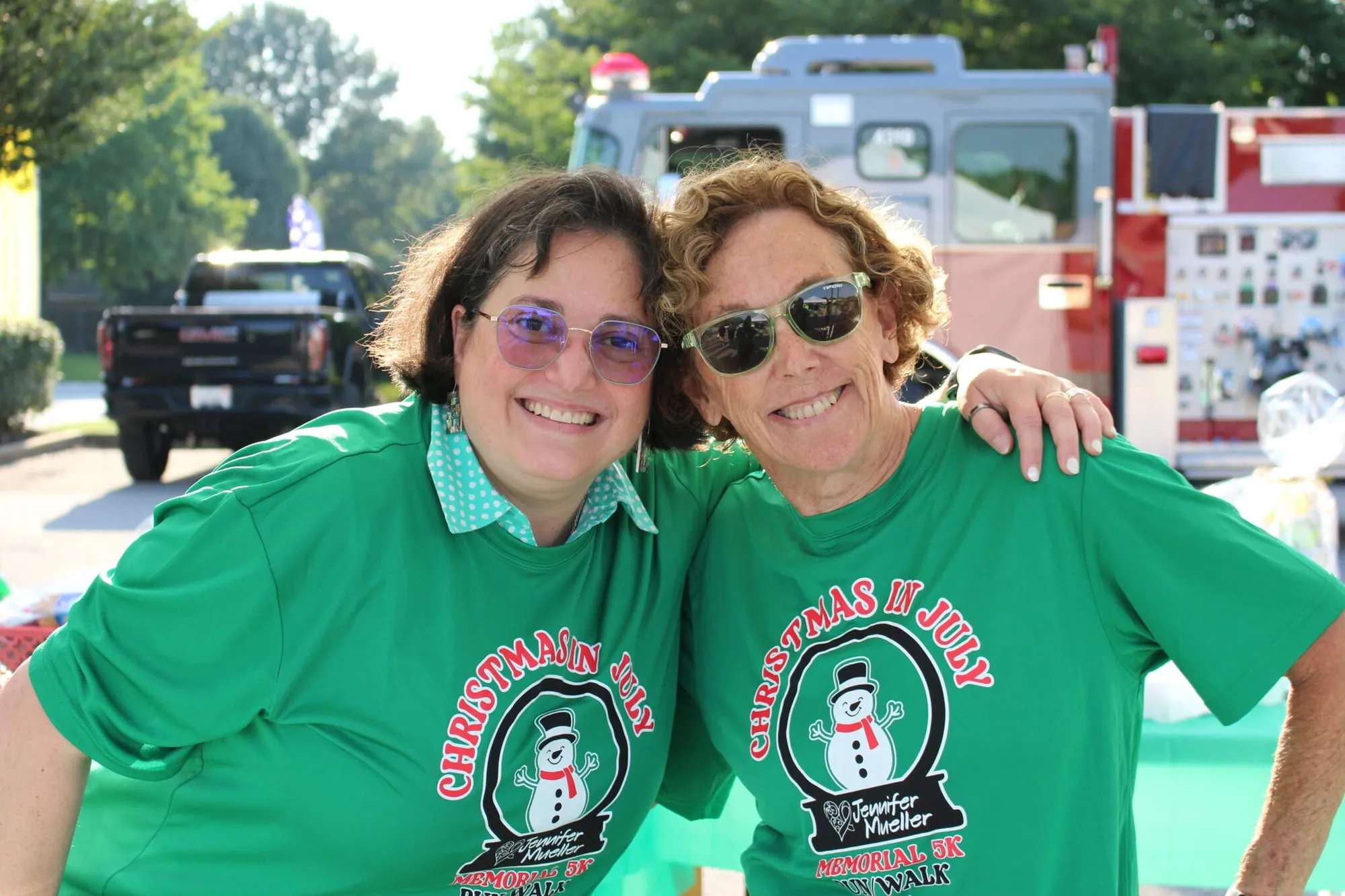 Two women in green “Christmas in July” race shirts smiling with their arms around each other.