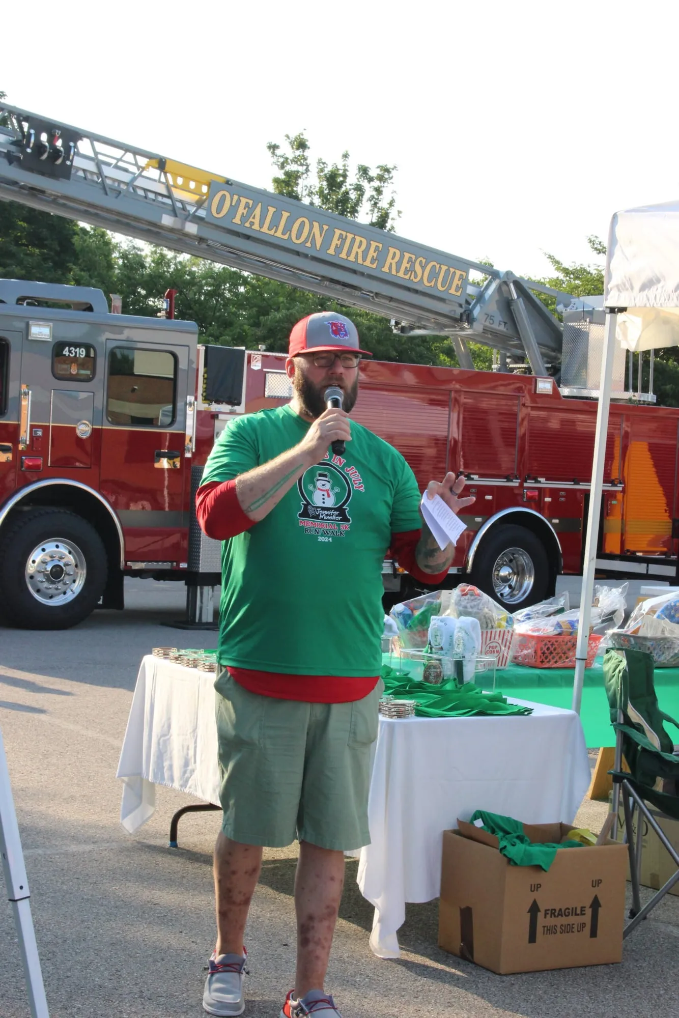 A man in a green race shirt speaking into a microphone beside a red fire engine.