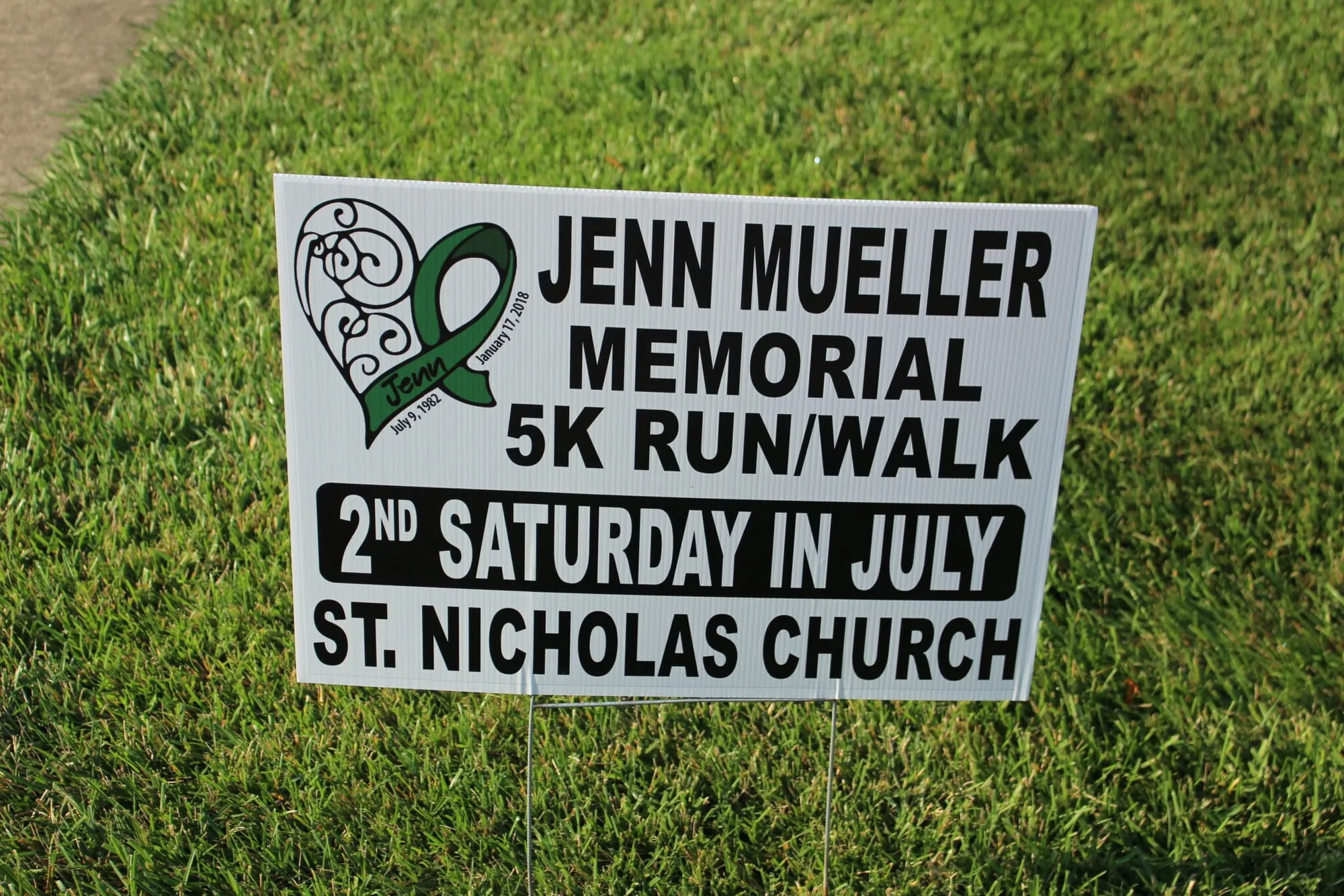 A roadside sign reading “Jenn Mueller Memorial 5K Run/Walk, 2nd Saturday in July, St. Nicholas Church”.