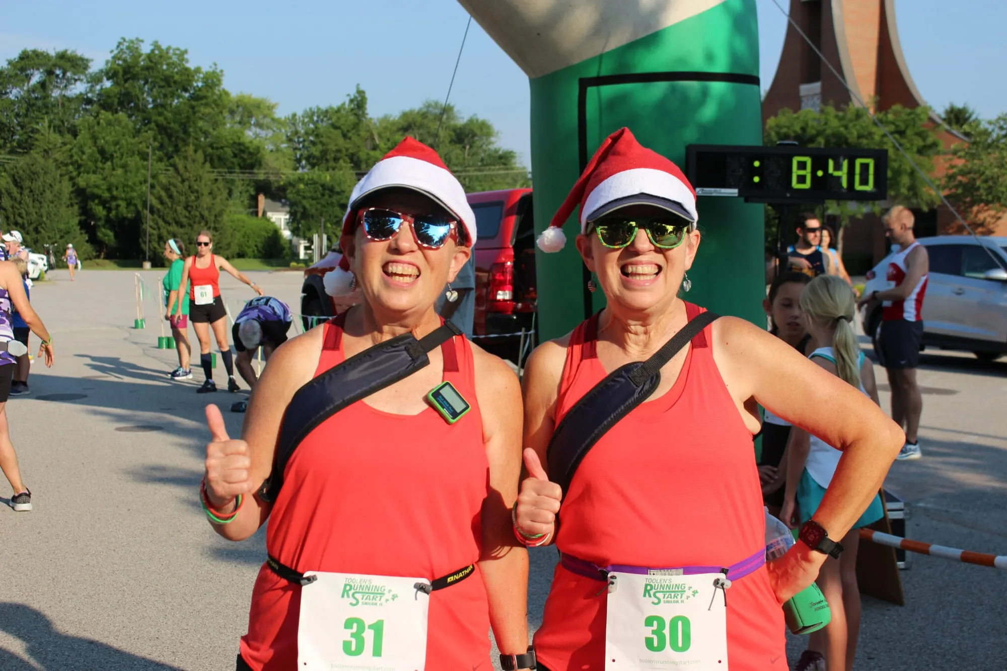 Two runners in Santa hats and red outfits posing in front of the race clock reading 8:40.